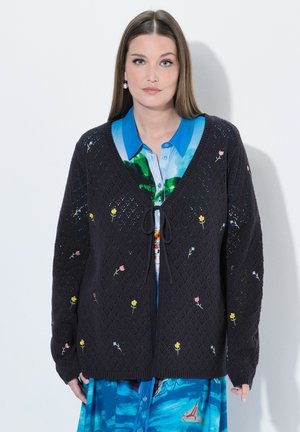 Vrouw met steil bruin haar draagt een zwarte gebreide cardigan met kleine geborduurde bloemen over een blauwe bloemenjurk met kraag en knopen.