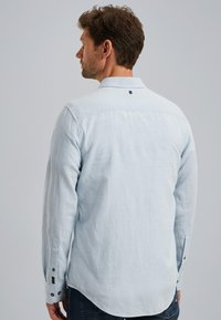 Camisa de manga larga azul claro con corte entallado, cuello con botones y dobladillo redondeado. Presenta botones en los puños y un pequeño detalle de logo.