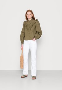Camisa de botões verde-oliva, com babados e mangas longas e volumosas; combinada com calças brancas de perna larga e uma bolsa de palha. As sandálias são verde-oliva.
