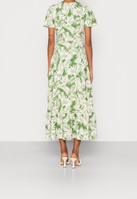 Woman wearing a green midi dress with white tropical leaf print, short sleeves, ruffled hem, and white heeled sandals, facing away.