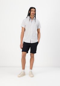 Camicetta a maniche corte con bottoni di colore bianco, con un motivo sottile, abbinata a pantaloni corti scuri arrotolati e scarpe da ginnastica chiare. Tessuto dalla consistenza liscia.