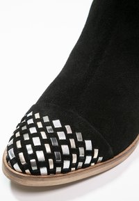 Punta de zapato de ante negro con acentos cuadrados metálicos plateados y negros tejidos en una suela de madera.