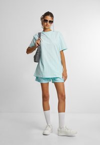 Világoskék, hasított oversized póló, függőleges csíkokkal, mintás rövidnadrággal párosítva. A modell egy szürke táskát tart, napszemüveget visel és fehér sportcipő van rajta.