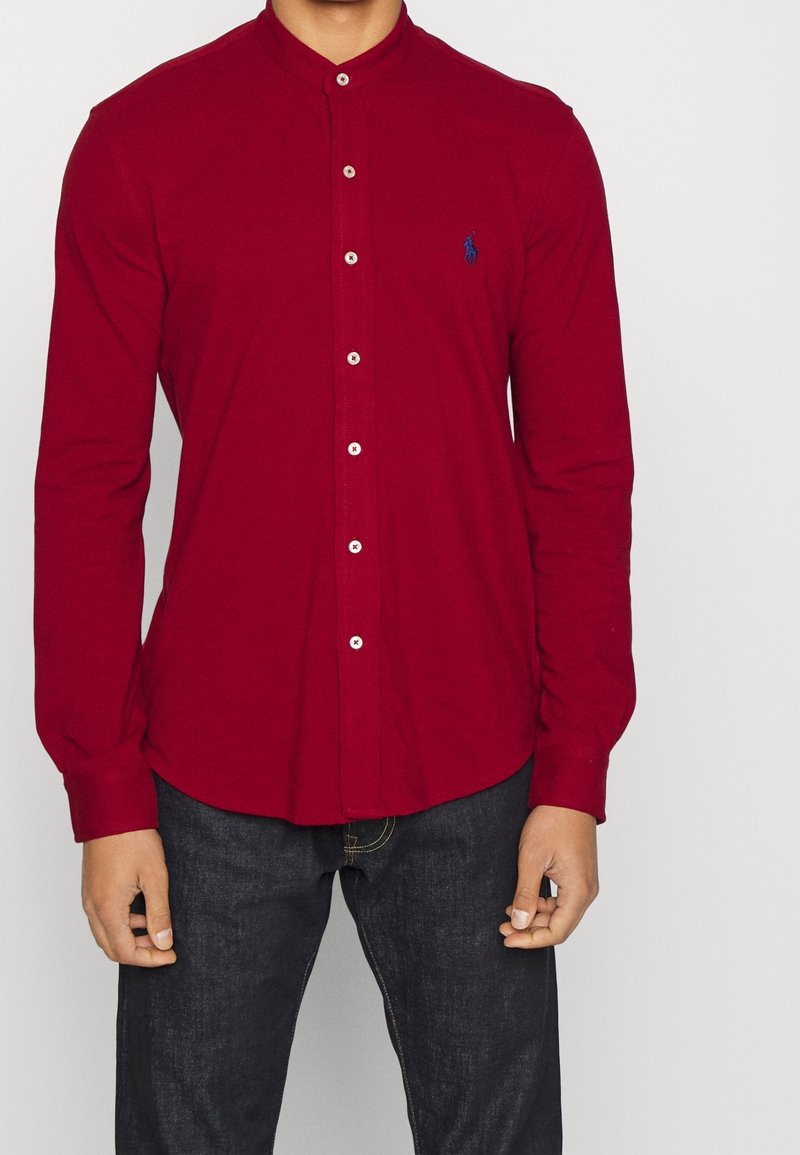 Homme portant une chemise rouge à boutons avec un petit logo bleu brodé sur la poitrine et un jean bleu foncé, debout devant un fond uni.
