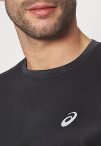 Schwarzes Sport-T-Shirt mit Rundhals, aus glattem Material. Mit einem weißen Logo auf der linken Brustseite.