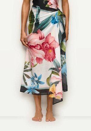 Femme portant une jupe sarong fleurie nouée avec de grandes fleurs roses, bleues et jaunes et des feuilles vertes, pieds nus sur fond blanc.
