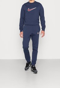 Ensemble de sweatshirt et pantalon Nike bleu marine, avec un logo en bordeaux et blanc. Accompagné de baskets noires, en tissu lisse, coupe ajustée.