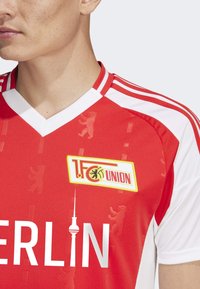 Maillot de sport rouge avec des accents blancs, présentant un col en V, un design texturé et un logo portant "1. FC Union" ainsi que la tour de télévision de Berlin.
