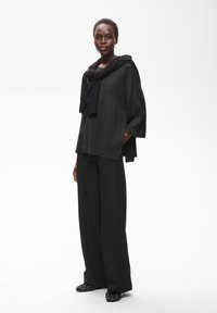 Femme debout portant un pull sombre ample avec un pull noir noué sur les épaules, un pantalon large noir et des chaussures noires sur fond blanc.