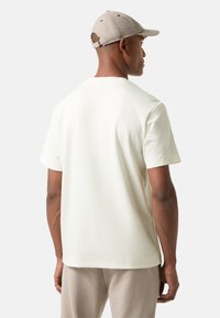 Lichtcrème t-shirt met korte mouwen, gemaakt van zachte stof, met een ontspannen pasvorm en een strakke uitstraling. gedragen met een beige pet.