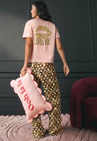 Rožinės spalvos medvilniniai marškinėliai su auksiniais užrašais nugaroje, raštuotos auksinės ir rudos pižaminės kelnės, laiko rožinę pagalvę su raudonais užrašais ir banguotu kraštu.