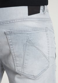 Jeans de color azul claro con un único bolsillo trasero que presenta un distintivo detalle bordado en forma de punta, complementado por un parche de cuero oscuro.