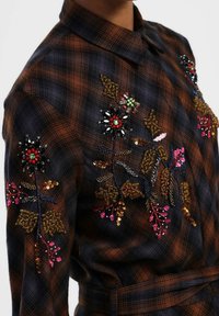 Camicia a quadri marrone e nera con modelli floreali ricamati che presentano paillettes e perline rosse, rosa, verdi e nere sulle maniche.