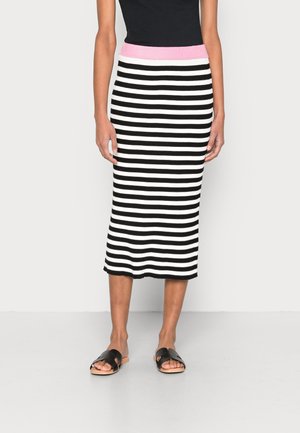 Femme portant une jupe midi à rayures horizontales noires et blanches avec une ceinture rose et des sandales slides noires, debout devant un fond uni.