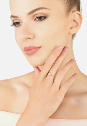 Gold ring with a minimal design featuring a 'C' accent and a small clear stone. Model's hand rests on her face, showcasing neutral nail polish.