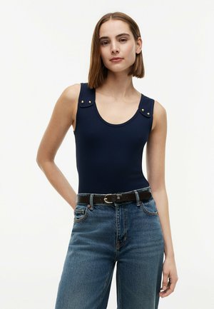Młoda kobieta o prostych brązowych włosach nosi granatowy, bezrękawnikowy prążkowany top z guzikami, wpuszczony w niebieskie, wysokiej talii jeansy i czarny pasek.