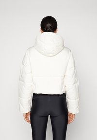 Bijela, skraćena puffer jakna s kapuljačom. Ima prošivene uzorke i široke rukave, uparen s crnim leggingsima. Glatka tekstura.