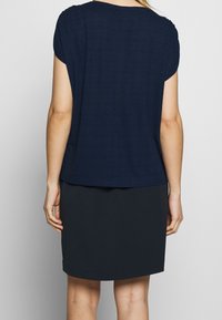Mujer vestida con una blusa de manga corta azul marino y una falda negra, de pie de espaldas a la cámara frente a un fondo blanco liso.