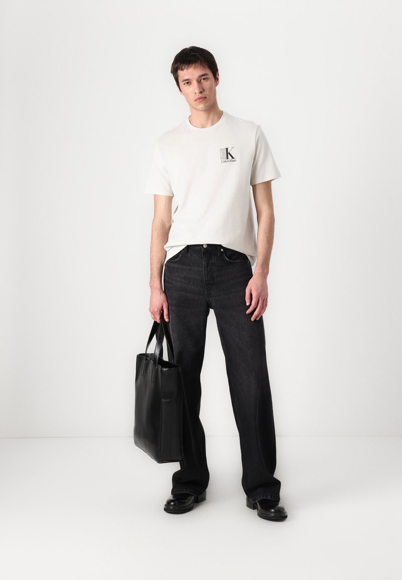 T-shirt blanche avec un logo, jeans en denim noir et un sac fourre-tout noir. Le modèle se tient sur un fond blanc, portant des chaussures noires.