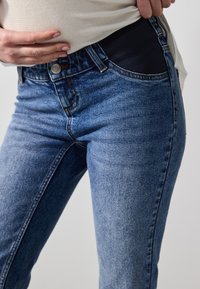 Kék farmernadrág gombos zárással, slim fit fazonnal és fekete gumis derékrésszel. Az anyag megjelenése elhalványult, texturált.