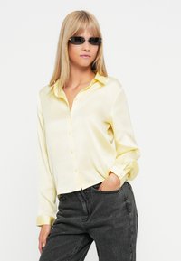 Jasnożółta satynowa koszula zapinana na guziki z kołnierzykiem, długimi rękawami i luźnym krojem; w połączeniu z ciemnoszarymi spodniami z wysokim stanem.