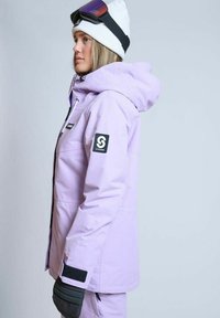 Lilla vinterjakke med hætte, lynlåslukning foran, justerbare snore og sorte velcro-ærmer. Inkluderer et logo-patch på ærmet.