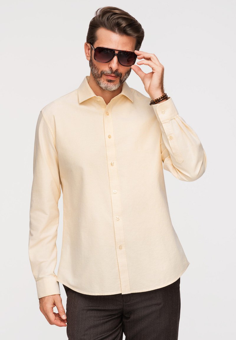 Homme avec une barbe portant une chemise beige boutonnée et des lunettes de soleil foncées, ajustant ses lunettes avec la main droite sur un fond uni.