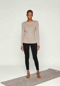 Strakke langemouwtop in lichtbeige katoen en zwarte leggings. Model staat op een gestructureerde grijze yogamat, blootsvoets, tegen een effen achtergrond.