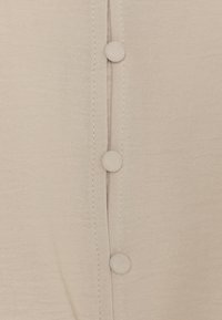Tessuto beige con tre bottoni foderati allineati verticalmente e cuciture visibili lungo la pattina dei bottoni.
