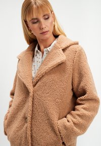 Kobieta z blond włosami, nosząca złote kolczyki w kształcie kół i tanowy, wzorzysty płaszcz teddy na białej bluzce w kropki, z rękami w kieszeniach płaszcza.