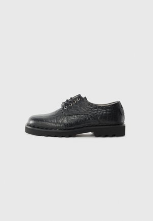 Zapato de vestir negro de cuero con un acabado texturizado, punta redonda y suela plana de goma con un patrón de tacos. Incluye ocho ojales para los cordones.