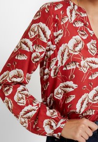 Rode blouse met bloemenprint in wit en bruin. Heeft lange mouwen, een V-halslijn en geplooide manchetten. Soepele stofstructuur.