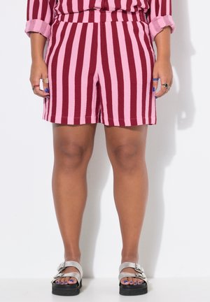 Benene på en person, der har røde og pink lodrette stribede shorts, sølvspænde sandaler, ringe og blå neglelak på tånegle og fingernegle.
