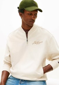 Felpa beige con mezza cerniera, polsini a coste e logo ricamato. Indossata sopra una camicia a righe. Cappellino verde con dettaglio logo.