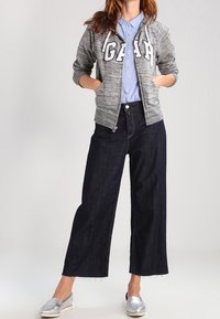 Felpa grigia con zip e lettering "GAP", camicia azzurro chiaro con bottoni, pantaloni scuri a gamba larga in denim e scarpe slip-on argento.