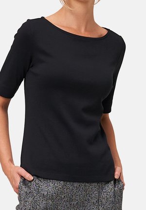 Femme portant un haut noir uni à manches courtes et un pantalon gris à motifs, les mains dans les poches, sur un fond clair.