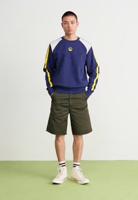 Námornícky sveter s žltými bočnými pruhmi, okrúhlym logom a bielymi akcentmi v kombinácii s olivovo zelenými šortkami a bielymi vysokými teniskami.