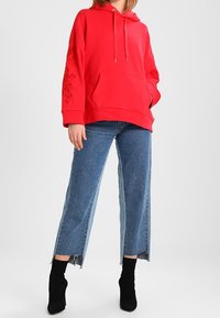 Sudadera con capucha roja con bolsillo delantero y estampado floral en las mangas, combinada con vaqueros de pierna ancha de color azul claro y botines negros.