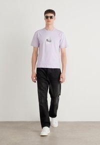 Light purple cotton t-shirt with a front print, paired with black denim jeans and white sneakers. Model wearing sunglasses, standing against a neutral background.