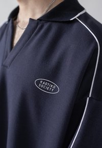 Mörkblå polotröja med krage, som har vit kantning längs axlarna och en cirkulär "KAOTIKO SOCIETY"-logotyp på bröstet. Slät textur.