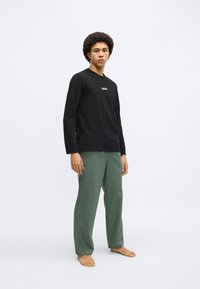 Cămașă neagră cu mâneci lungi și logo-ul "BOSS"; pantaloni verzi în carouri; croială lejeră; fundal simplu; modelul este desculț.