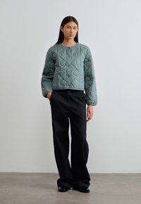 Πράσινο καπιτονέ μπουφάν με στρογγυλή λαιμόκοψη και μακριά μανίκια, σε συνδυασμό με μαύρο παντελόνι φαρδιάς γραμμής και μαύρα παπούτσια σε ουδέτερο φόντο.