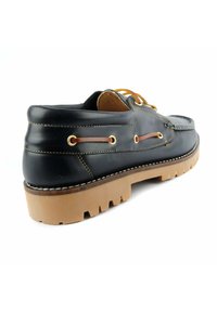 Zapato de barco de cuero negro con perfil bajo, suela de goma y cordones en color marrón. Presenta ojales dorados, costuras en contraste y una superficie texturizada.