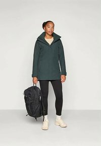 Tmavozelená nepriepustná bunda s vysokým golierom, skombinovaná s čiernymi legínami a čiernym ruksakom, nosená na béžovom topu. Svetlé tenisky.