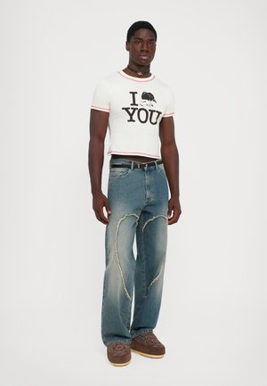Hombre de pie con una camiseta blanca cortada que dice "Te [cangrejo]" , jeans azules con parches desgastados en forma de corazón y zapatos marrón con un patrón.