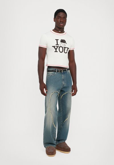 Mann steht da und trägt ein weißes, kurzes T-Shirt mit dem Aufdruck "I [Krabbe] YOU", blaue Jeans mit herzförmigen, ausgefransten Patches und braunen gemusterten Schuhen.