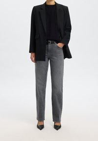 Personne portant un blazer noir, un top noir, un jean gris droit avec une ceinture noire et des chaussures noires à bout pointu, debout avec une main dans la poche.