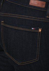 Jeans in denim scuro con cuciture gialle a contrasto, con un'etichetta in pelle con la scritta "Tommy Hilfiger" sulla cintura e un piccolo dettaglio con la bandiera tricolore sulla tasca.