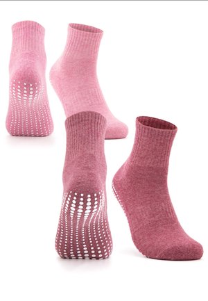 Füße mit bordeauxfarbenen rutschsicheren Grip-Socken mit weißen gepunkteten Traktionslinien an den Sohlen, kombiniert mit schwarzen Leggings vor weißem Hintergrund.