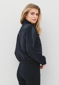 Fiatal nő szőke hajjal, fekete mintás ujjú dzsekit és fekete nadrágot visel, hátrafordulva néz a vállán túlra.